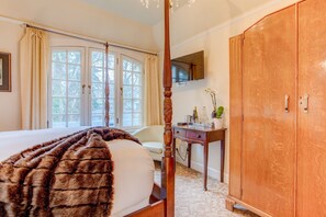 A bedroom with a four-poster bed, a wooden wardrobe, a desk with a vase, and a television on the wall.