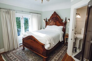 Une chambre à coucher avec un grand lit en bois, une fenêtre avec des rideaux, une salle de bain avec un lavabo et un miroir, et un tapis à motifs.