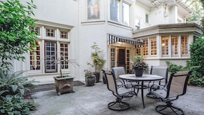 A courtyard with a round table and chairs, surrounded by potted plants and a white building with large windows.