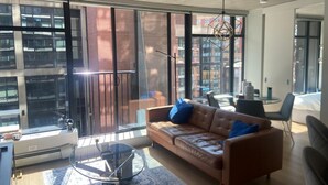 Una sala de estar moderna con un sofá de cuero, una mesa de centro de vidrio y ventanas amplias con vistas a un paisaje urbano.