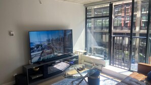 Una sala moderna con un televisor de pantalla plana grande, una mesa de centro de vidrio, un sofá y vistas de un paisaje urbano a través de amplios ventanales.