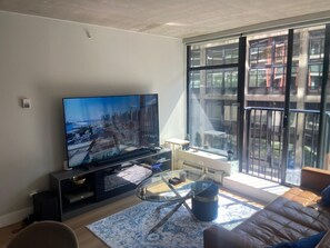 Een moderne woonkamer met een grote flatscreen televisie, een glazen salontafel, een bank en een uitzicht op een stadsgezicht door grote ramen.