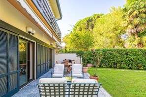 A patio with outdoor furniture, a glass door entrance, and a well-maintained garden with a hedge and trees.