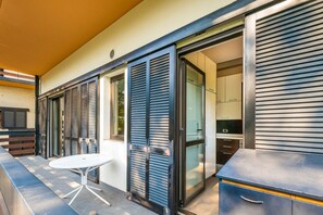 Une terrasse extérieure moderne, dotée d’une table et de chaises, d’une porte-fenêtre coulissante donnant sur une cuisine et d’un volet roulant fermé.