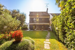 Une maison à deux étages avec un balcon, un jardin avec une pelouse et des plantes en pot, et un chemin de pierre menant à l’entrée.