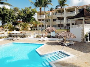 A pool area with lounge chairs, umbrellas, and a building with balconies. - Beautiful 1-bdrm suite steps from the beach in Puerto Plata with deck and AC (Puerto Plata)