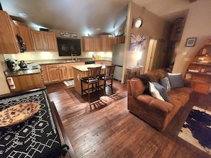 A kitchen with wooden cabinets, a dining area with chairs, a sofa, and a rug.