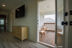 Et værelse med trægulv, en trækommode, et fladskærms-tv og en skydedør af glas, der fører til en udendørs balkon med fletstole.