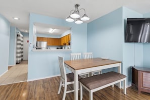 A dining area with a wooden table and chairs, a kitchen in the background, and a flat-screen TV mounted on the wall. - Sea Oats J206 Large 2 bedroom 2 bath Pool,Waterfront,Beach access,Pet friendly (Gulf Shores)