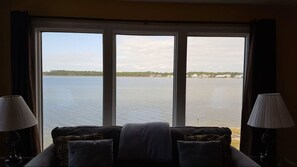 Ein Zimmer mit großem Fenster mit Blick auf ein Gewässer, einem Sofa mit Kissen und je einer Lampe auf beiden Seiten.