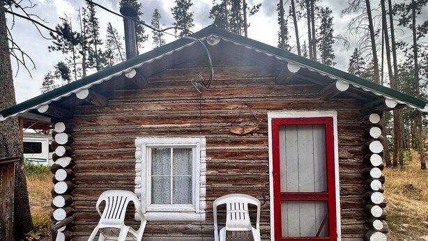 A log cabin with two white plastic chairs outside, a red door, and a window. - Cabin #3 Cozy one room cabin in charming Gould, perfect for a getaway. (Walden)