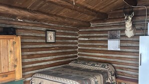 A log cabin interior with a bed, wooden walls, a ceiling fan, a mounted animal head, and a small table. - Cabin #3 Cozy one room cabin in charming Gould, perfect for a getaway. (Walden)