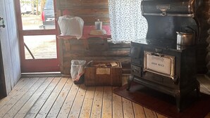 A rustic cabin interior with a wooden floor, a stove, a door leading outside, and a small table with a checkered tablecloth. - Cabin #3 Cozy one room cabin in charming Gould, perfect for a getaway. (Walden)