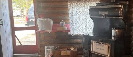 A rustic cabin interior with a wooden floor, a stove, a door leading outside, and a small table with a checkered tablecloth.