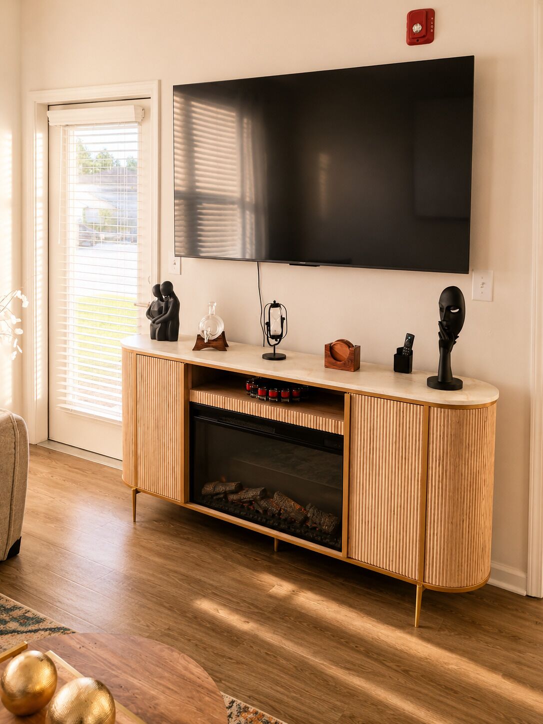 Un salon moderne doté d’un grand téléviseur à écran plat fixé au mur, d’un meuble de rangement en bois intégrant une cheminée, et d’une porte-fenêtre coulissante donnant sur une terrasse.