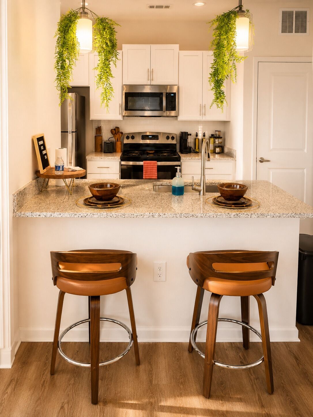 A kitchen with white cabinets, stainless steel appliances, a granite countertop, two wooden bar stools, and hanging plants.