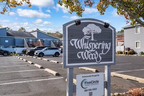 Schild für Whispering Wood mit Baumlogo, Parkplatz und Gebäuden im Hintergrund.