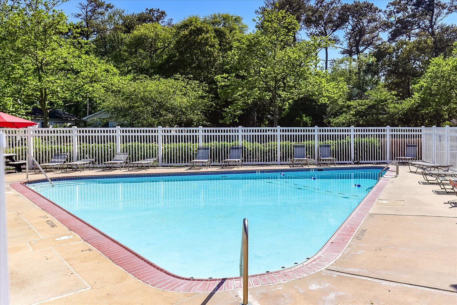 A rectangular swimming pool with a white fence, surrounded by lounge chairs and a red umbrella.