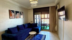 Ein Wohnzimmer mit einem blauen Sofa, einem kleinen runden Tisch, einem gemusterten Teppich, einer Chandelierleuchtre und einem an der Wand montierten Fernseher.
