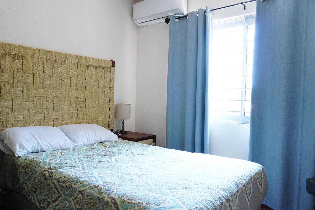 A room with a woven headboard, a bed with patterned bedding, a nightstand with a lamp, and a window with curtains.