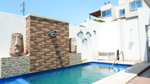 A swimming pool with a stone wall and shark sculptures, a metal ladder, and a white building with balconies. - Bungalows Huerta del Mar (Santa Cruz)