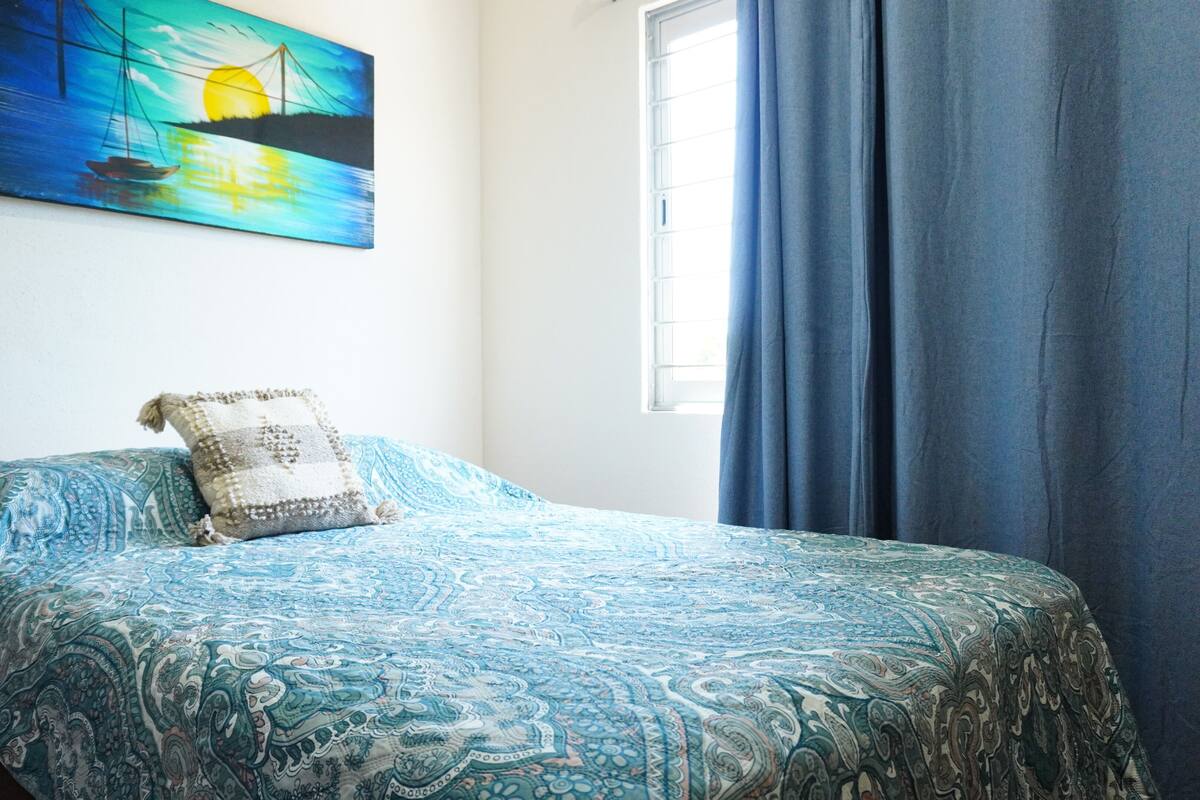 A bed with a patterned bedspread, a pillow, a painting on the wall, and a window with curtains.