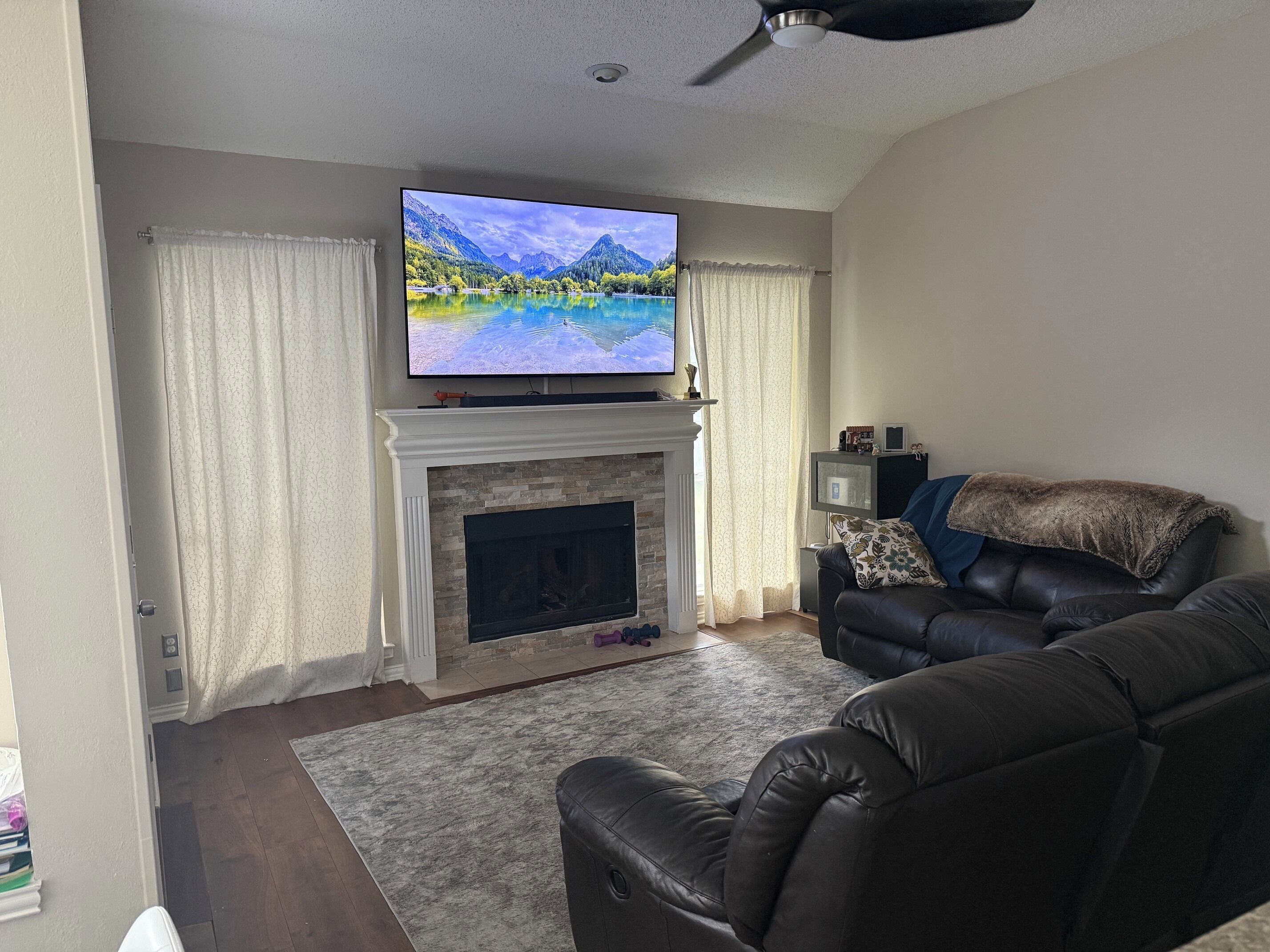 A living room with a fireplace, a flat-screen TV mounted above it, a leather sofa, and a ceiling fan.