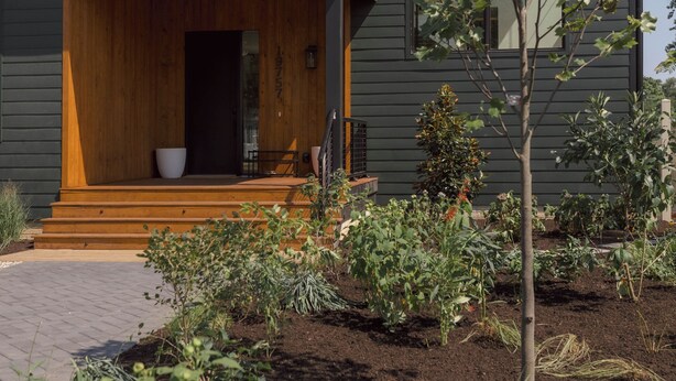 Ein modernes, zweistöckiges Haus mit einer hölzernen Veranda, einem kleinen Garten und einem Baum davor.
