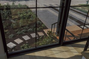 Blick durch ein Fenster auf einen Gartenbereich mit trittsteinförmigen Wegen, einer Holzdielen-Terrasse und einer Metalltreppe.