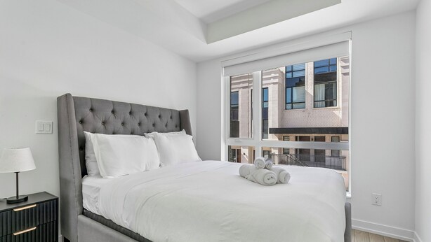 A modern bedroom with a grey tufted headboard, white bedding, a bedside table with a lamp, and a view of a building through a large window. - Home Richmond Hill 2bed2bath | Parking (Richmond Hill)