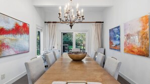 Ein Esszimmer mit einem langen Holztisch, grauen Polsterstühlen, einer Chandelierleuchtre und abstrakten Gemälden an den Wänden.