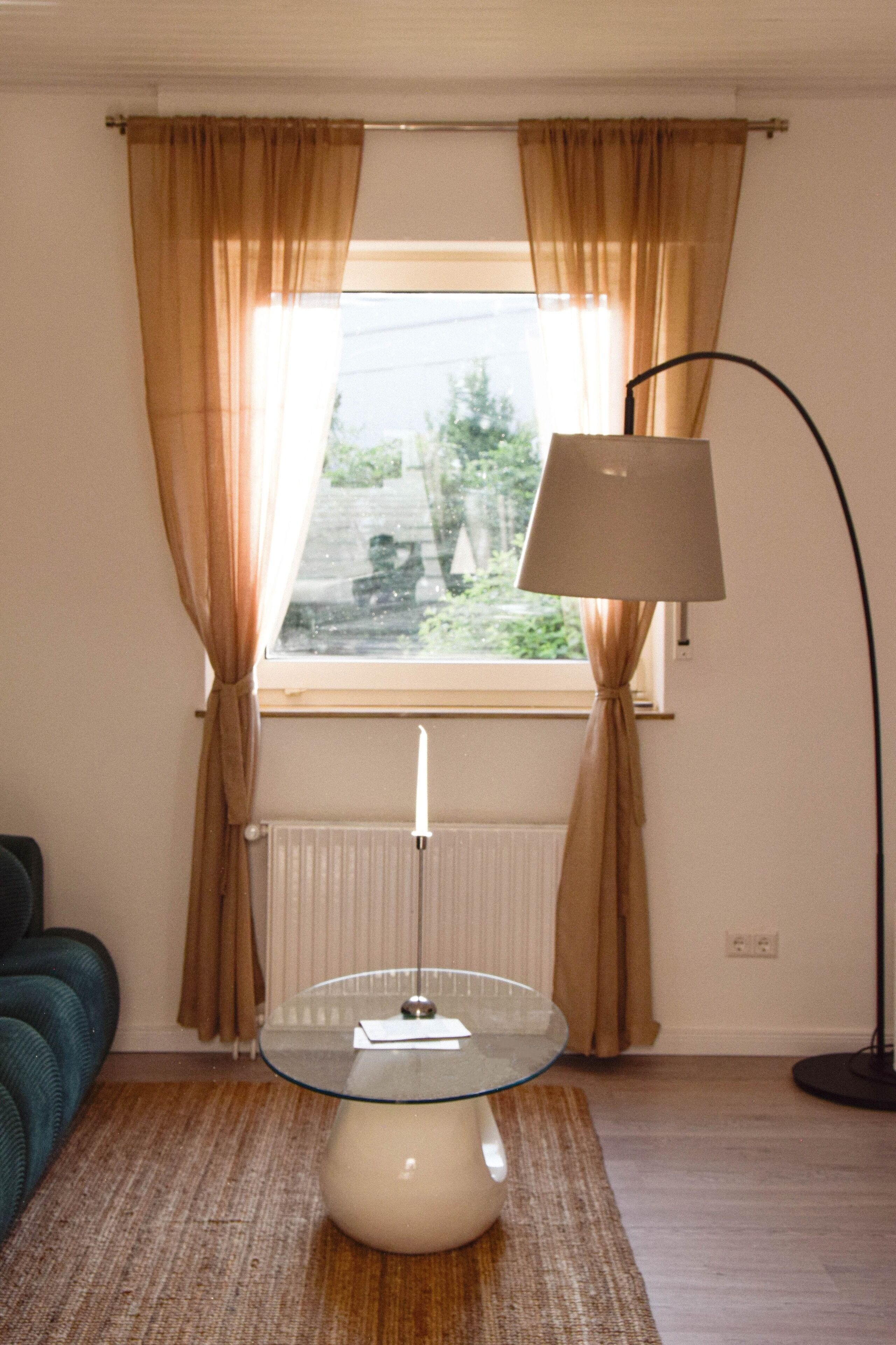Una habitación con una mesa de cristal, una lámpara de pie, un radiador y una ventana con cortinas.