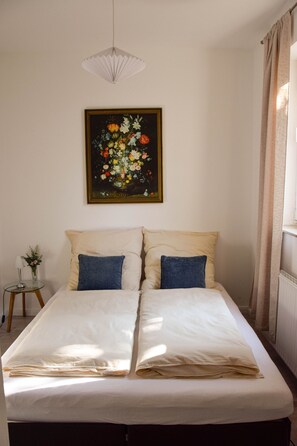 A double bed with two pillows, a painting of flowers on the wall, a small table with a vase, and a hanging light fixture.