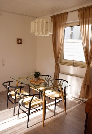 A dining area with a glass table, four chairs, a hanging light fixture, and a window with curtains.