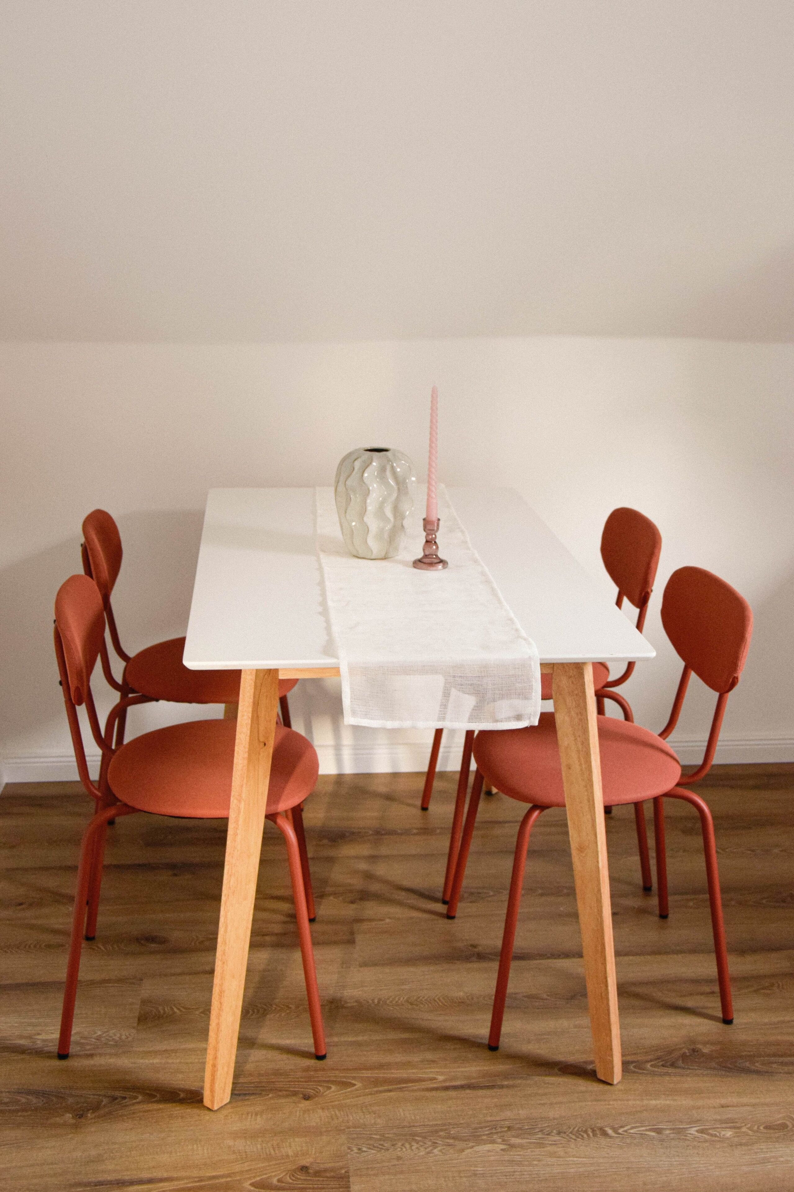 Une table à manger recouverte d’une nappe blanche, six chaises rouges, un vase et une bougie rose.