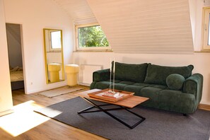 Ein Zimmer mit einem grünen Sofa, einem Holztisch, einem Spiegel und einem Fenster mit Jalousien.