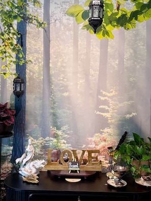 A room with a forest-themed backdrop, a decorative word 'LOVE' sign, a small statue, and a table with a teapot and a cup.