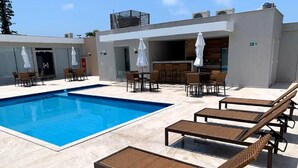 Una piscina con tumbonas y sombrillas, un área de bar y la fachada moderna de un edificio.