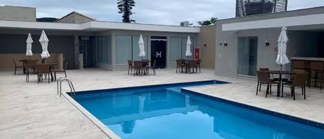 A swimming pool with surrounding tables and chairs, a covered seating area, and a building with a modern design.