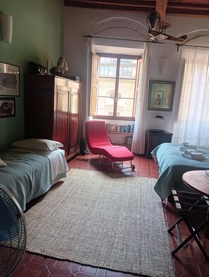 A room with two beds, a red chair, a small table, a fan, and a window with curtains. - Florentine Renaissance, to experience authentic Florence in the heart of the city (Firenze)
