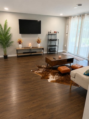Una sala de estar con televisor de pantalla plana, una mesa de centro de madera, otomanas de cuero, una planta en maceta y una alfombra.