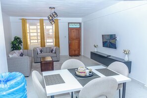 A modern living room with a sofa, dining table, and chairs, a wooden door, and a painting on the wall. - Shanvinhomes - 2 Bedroom Apartment in Elgonview near Reale Hospital (Eldoret)