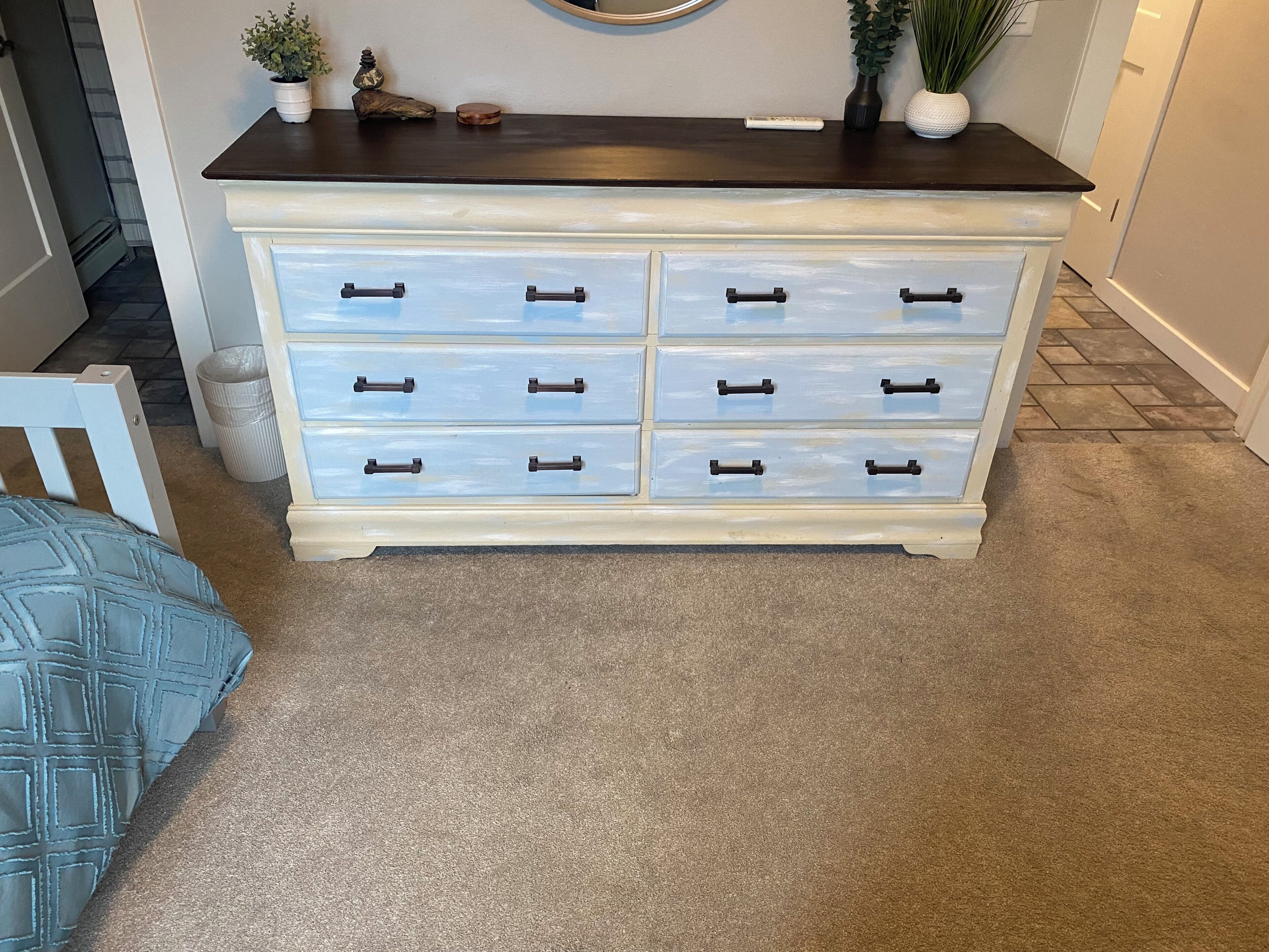 A white dresser with six drawers and black handles, placed against a wall with a mirror and decorative items.