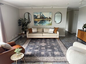 Een woonkamer met een bank, een salontafel en een vloerkleed. Aan de muur hangt een schilderij en er staat een plant aan de zijkant.