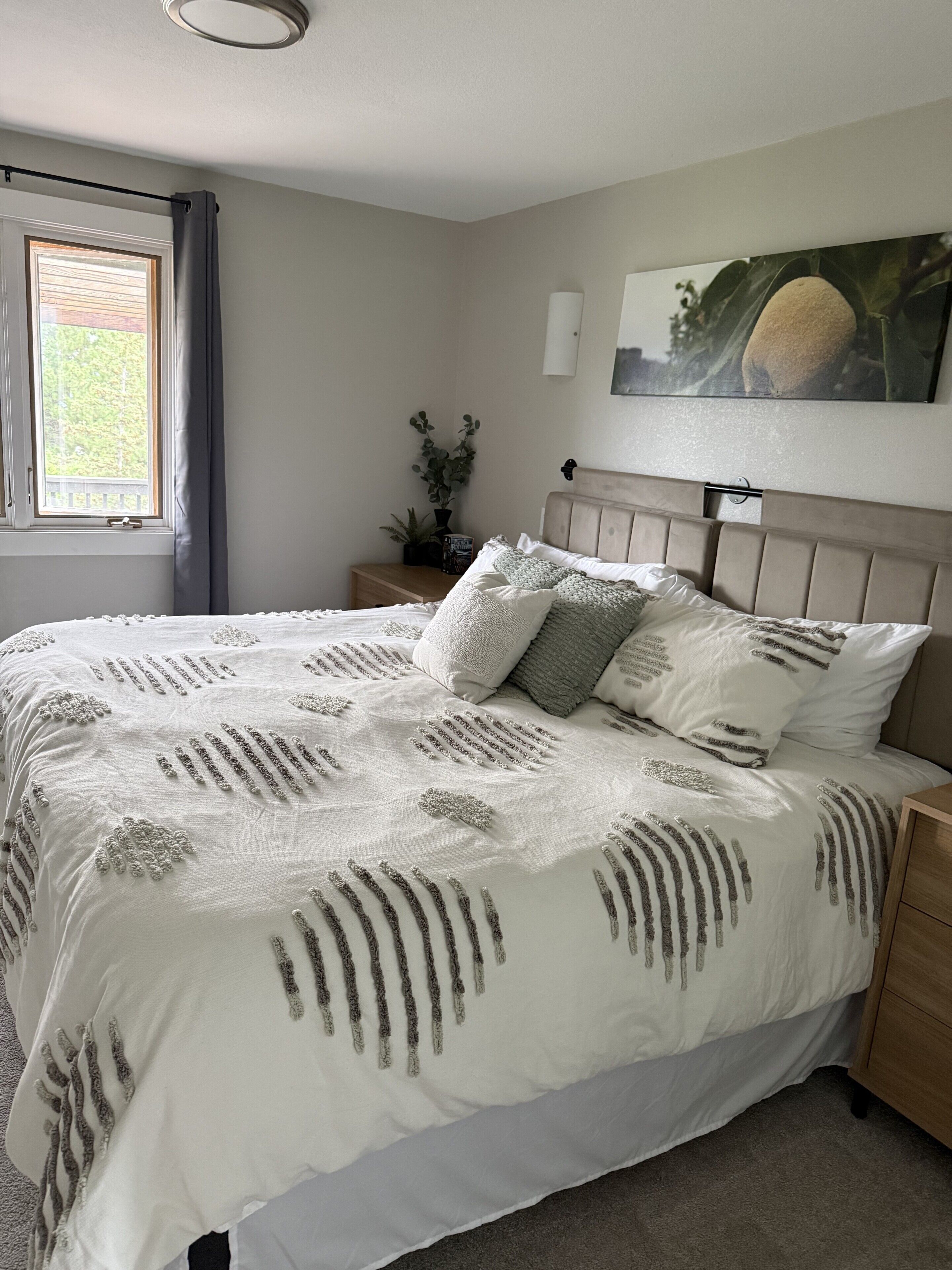A bedroom with a large bed, a nightstand, a window with curtains, a wall-mounted light, and a painting.