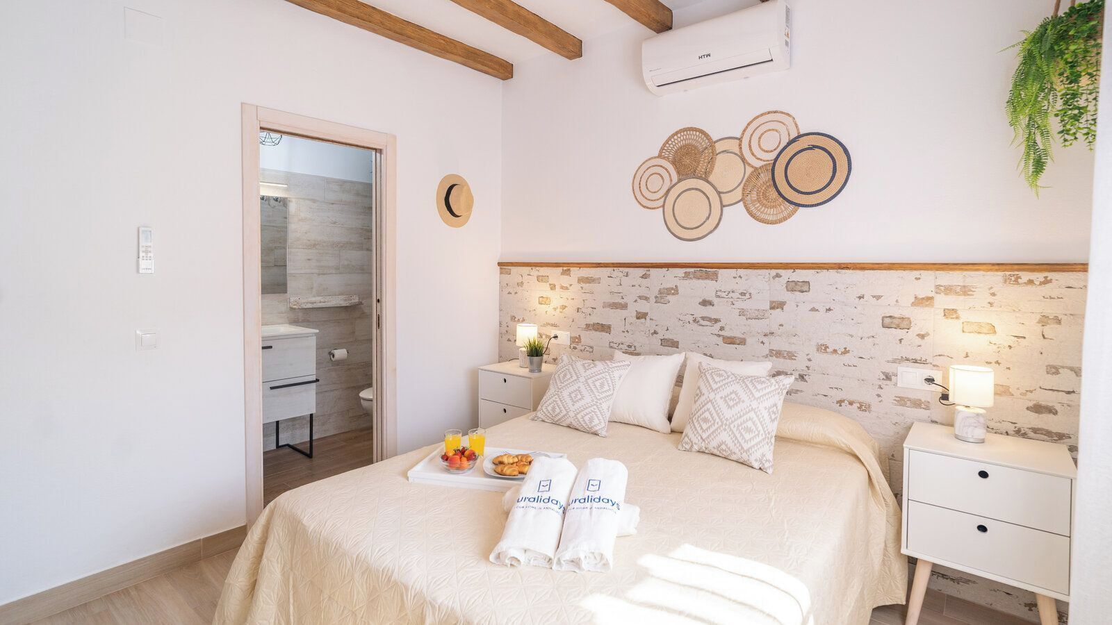 A hotel room with a bed, bedside tables, a wall clock, a wall-mounted air conditioner, and a small plant.