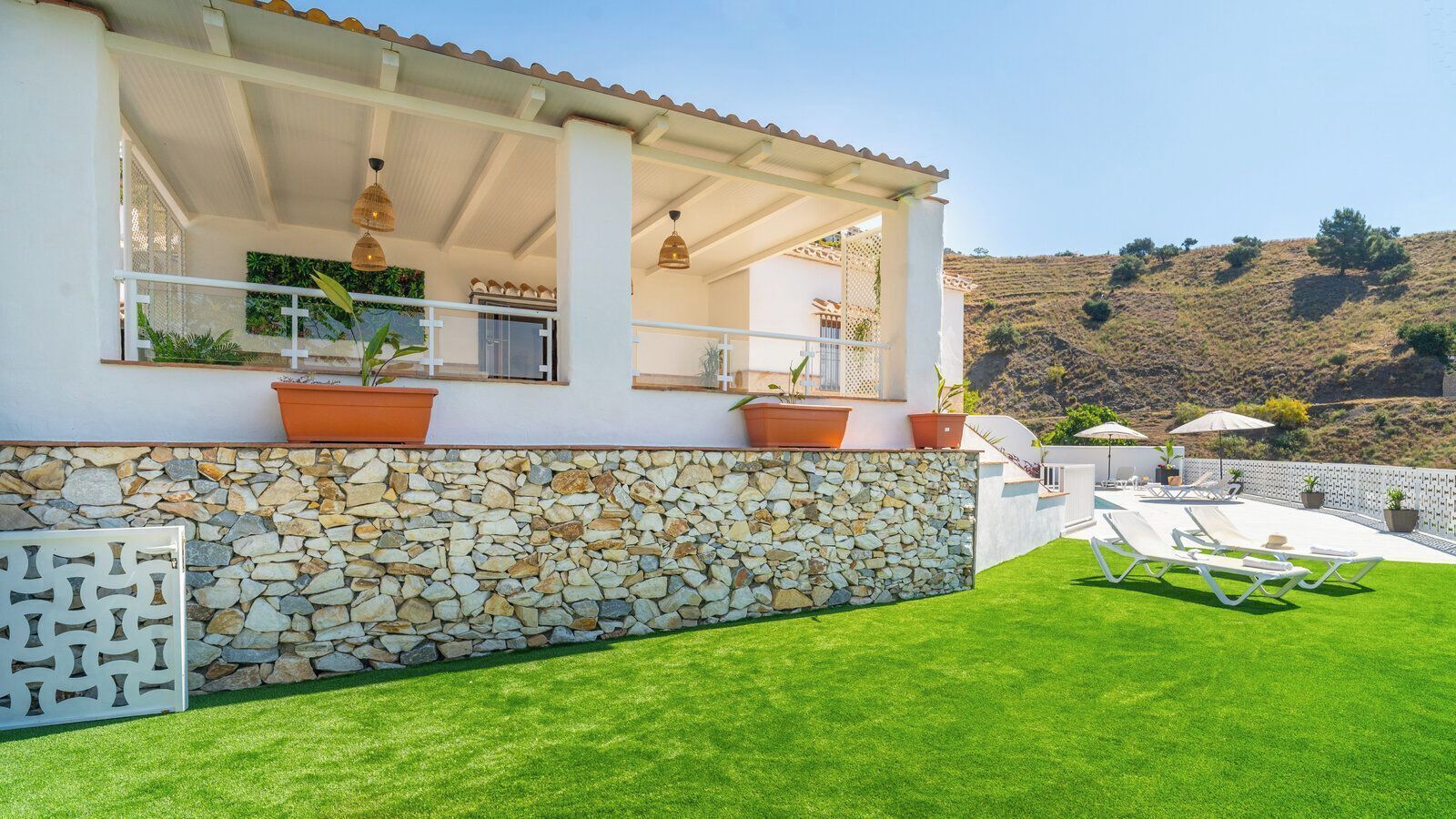 A house with a stone wall, a covered patio with outdoor furniture, and a view of a hilly landscape.