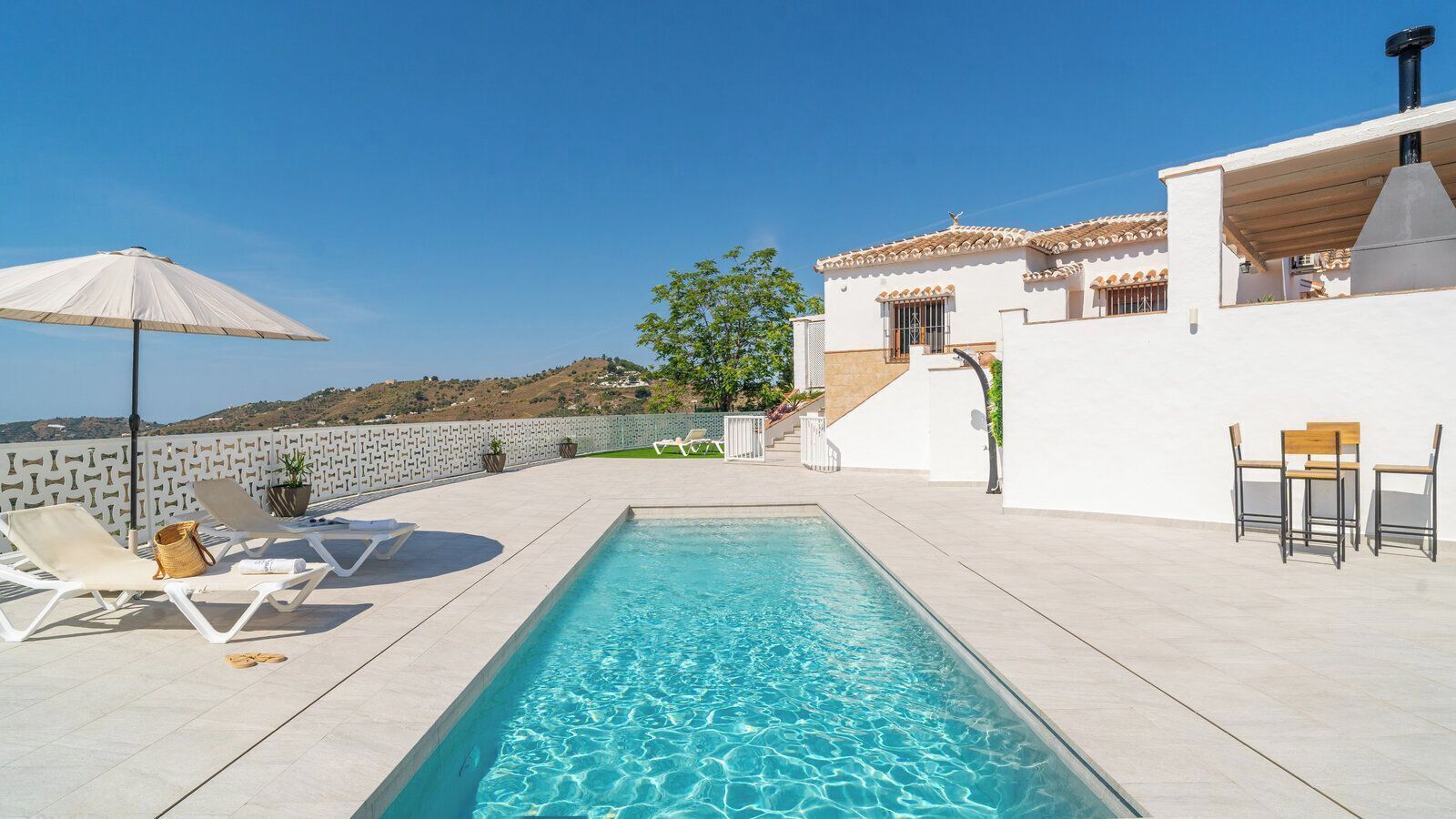 A swimming pool with lounge chairs and a table, surrounded by a white building with a tiled roof.