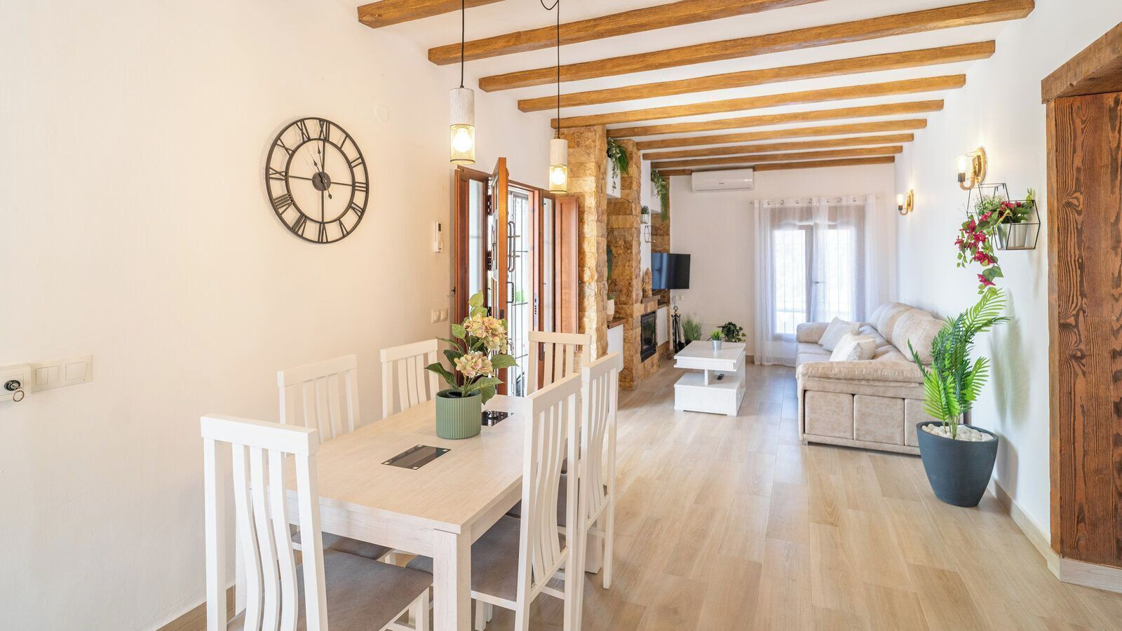 A dining area with a wooden table and chairs, a clock, and a living room with a sofa and a potted plant.