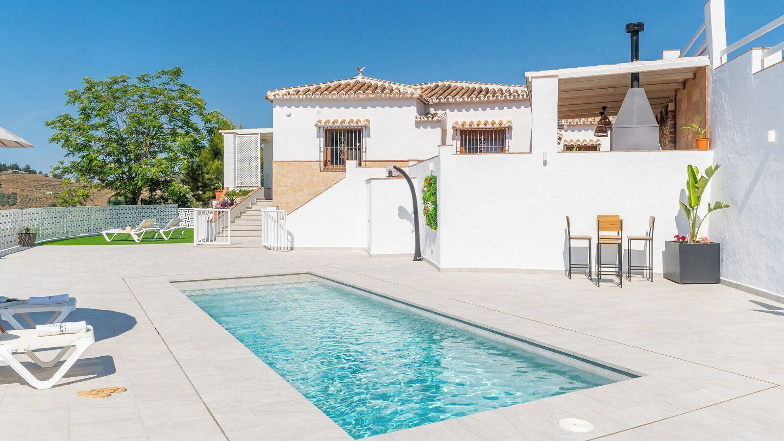 Una piscina con sillas de descanso, un edificio blanco con tejado de baldosas y una escalera que conduce a una zona cubierta.